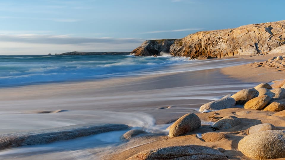A beach with smooth sand, large rocks, and a rocky cliff.