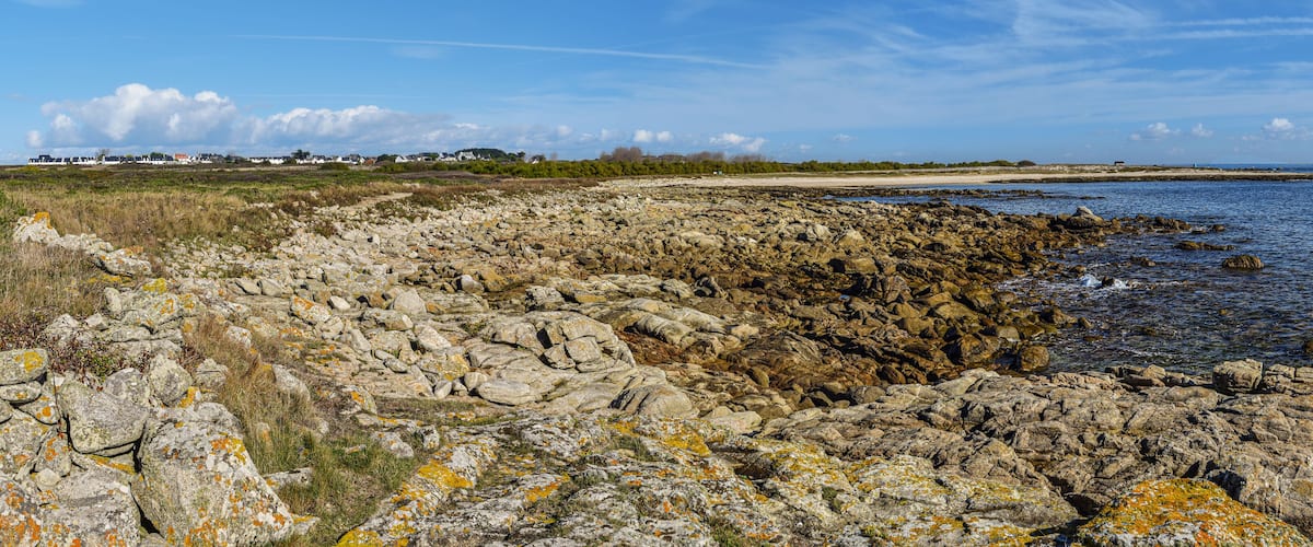 A rocky shoreline with a clear view of the sea, a sandy beach, and distant houses.