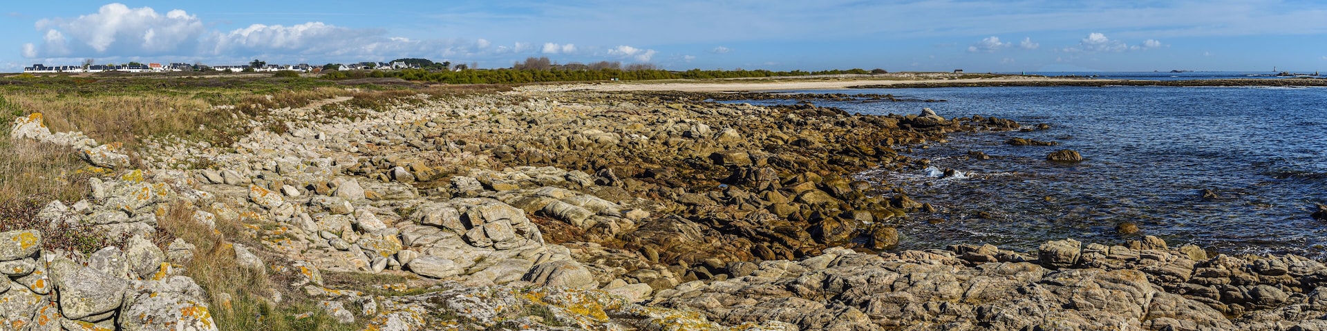 A rocky shoreline with a clear view of the sea, a sandy beach, and distant houses.