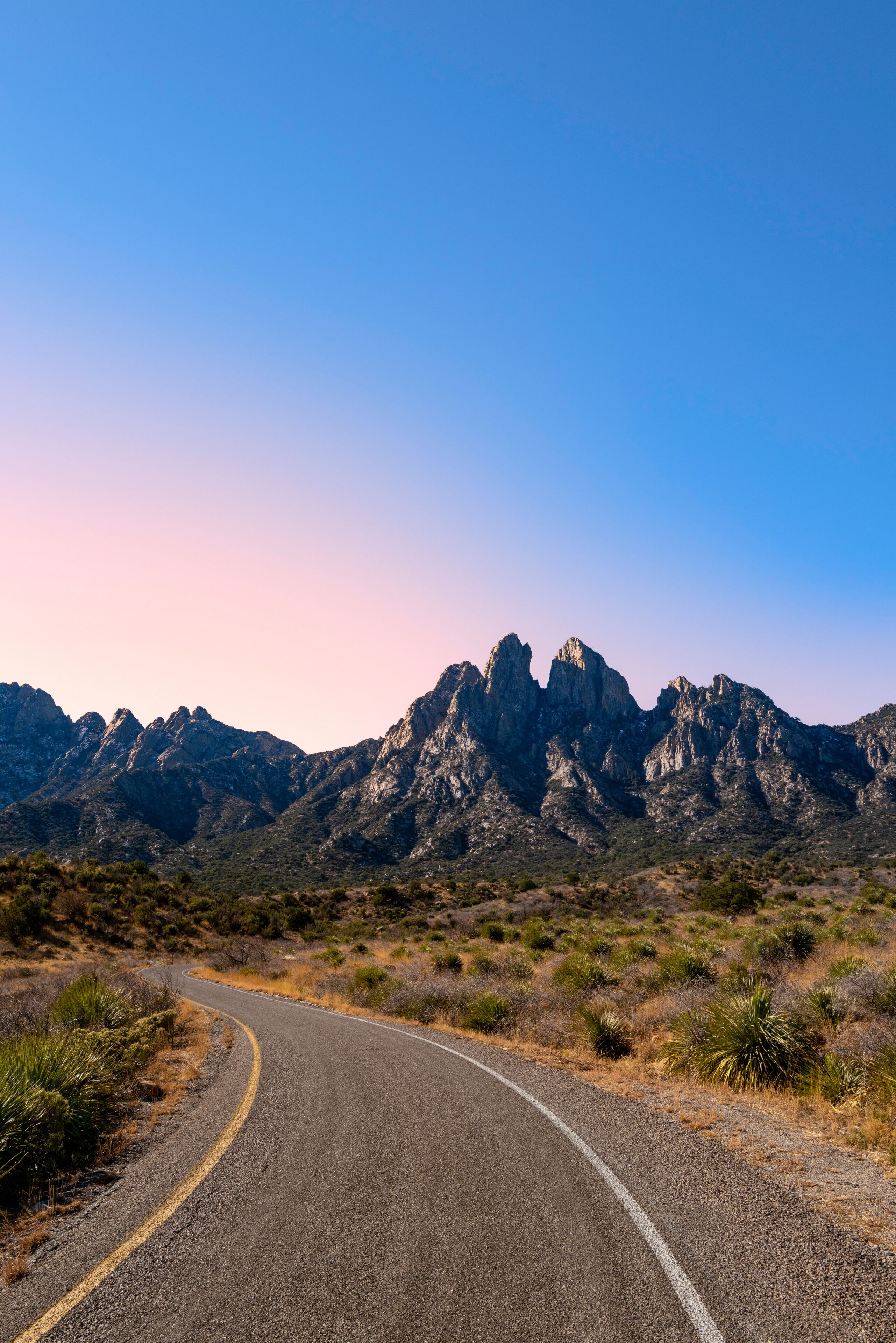 A winding road in a desert landscape with rocky mountains in the background.
