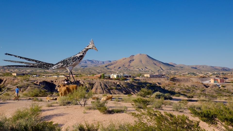 A desert landscape with a large bird sculpture, a mountain, and sparse vegetation.