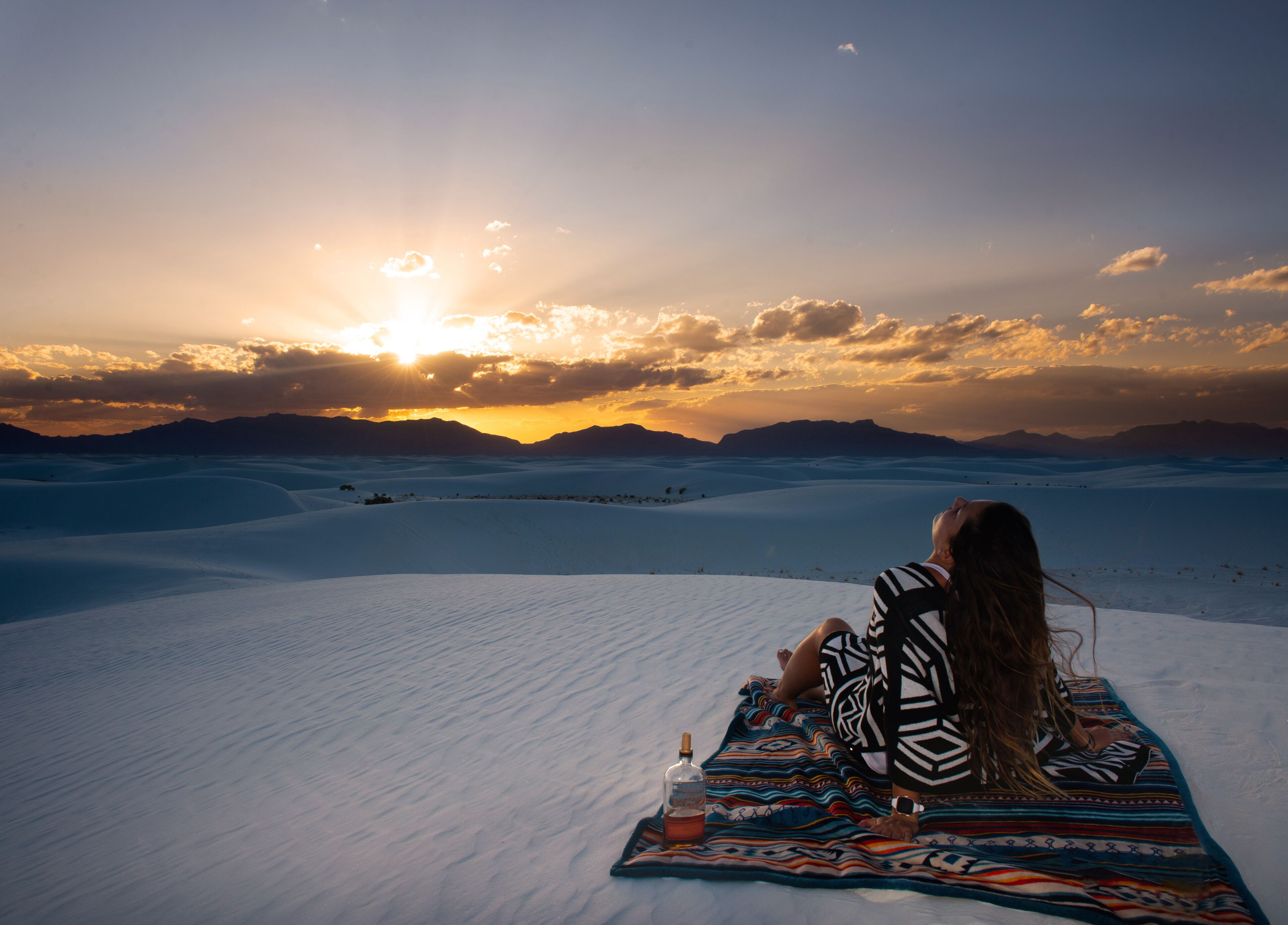 Uma pessoa relaxando em um cobertor em uma paisagem desértica com dunas de areia e um pôr do sol.
