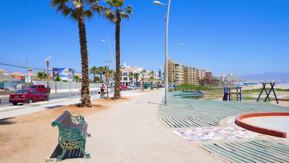 A coastal promenade with palm trees, a sandy path, a bench, a playground, and a hotel in the background.