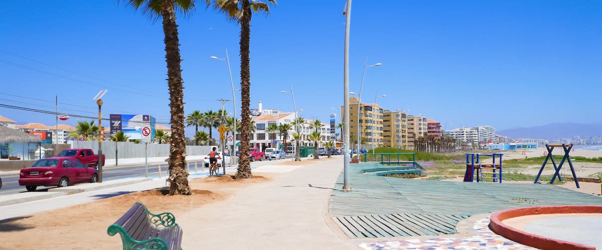 A coastal promenade with palm trees, a sandy path, a bench, a playground, and a hotel in the background.