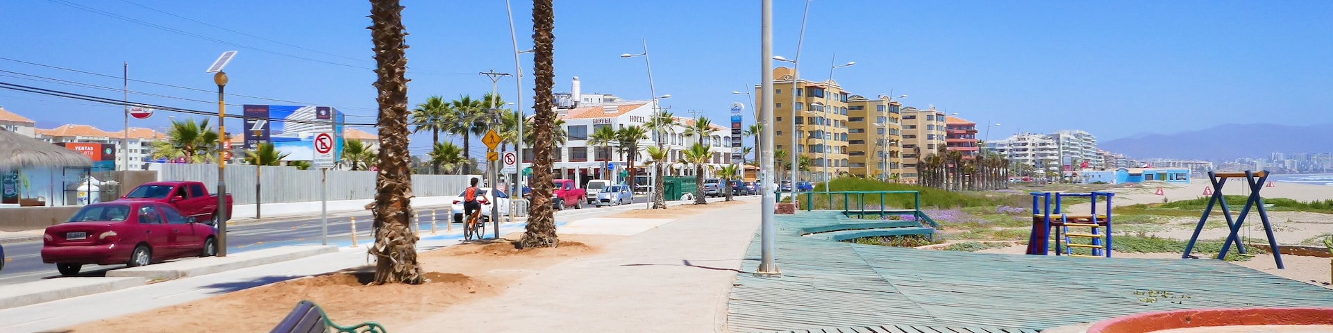 A coastal promenade with palm trees, a sandy path, a bench, a playground, and a hotel in the background.