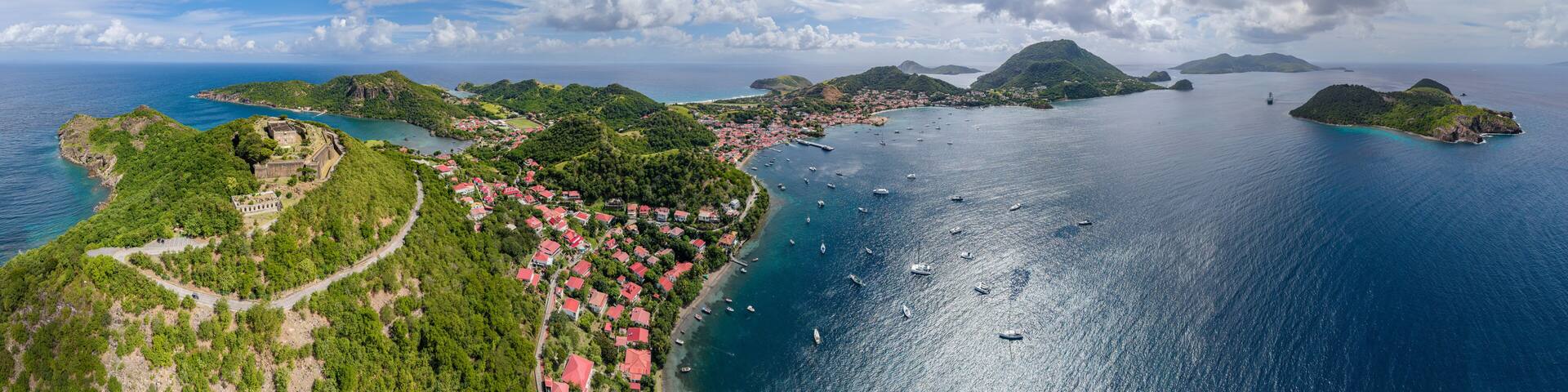 An aerial view of a coastal town with red-roofed houses, a fort, and a clear blue sea with boats.
