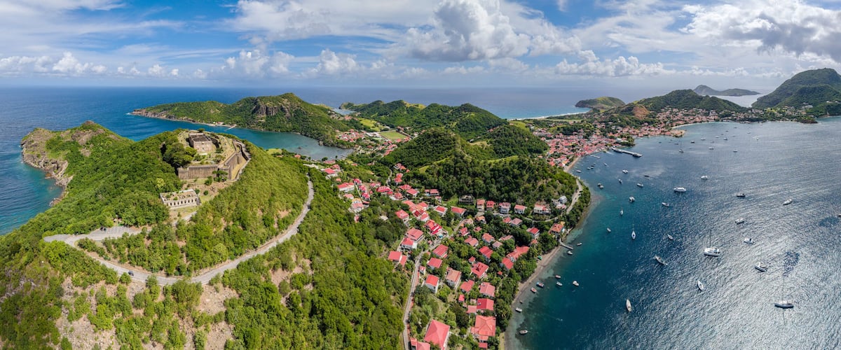 An aerial view of a coastal town with red-roofed houses, a fort, and a clear blue sea with boats.