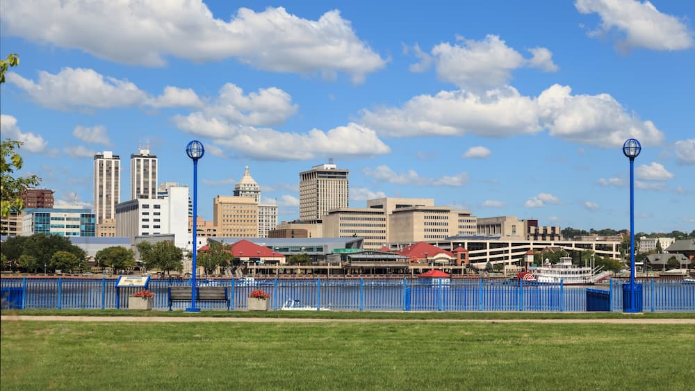 A city skyline with tall buildings, a river, and a blue fence in the foreground.