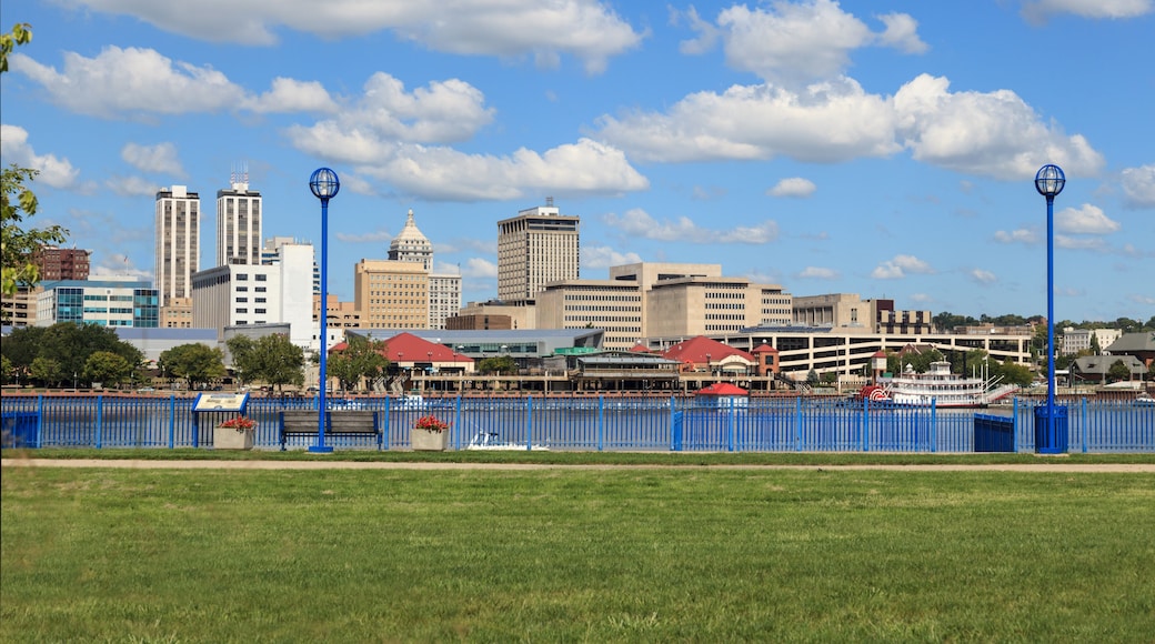 A city skyline with tall buildings, a river, and a blue fence in the foreground.