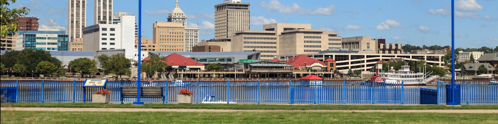 A city skyline with tall buildings, a river, and a blue fence in the foreground.