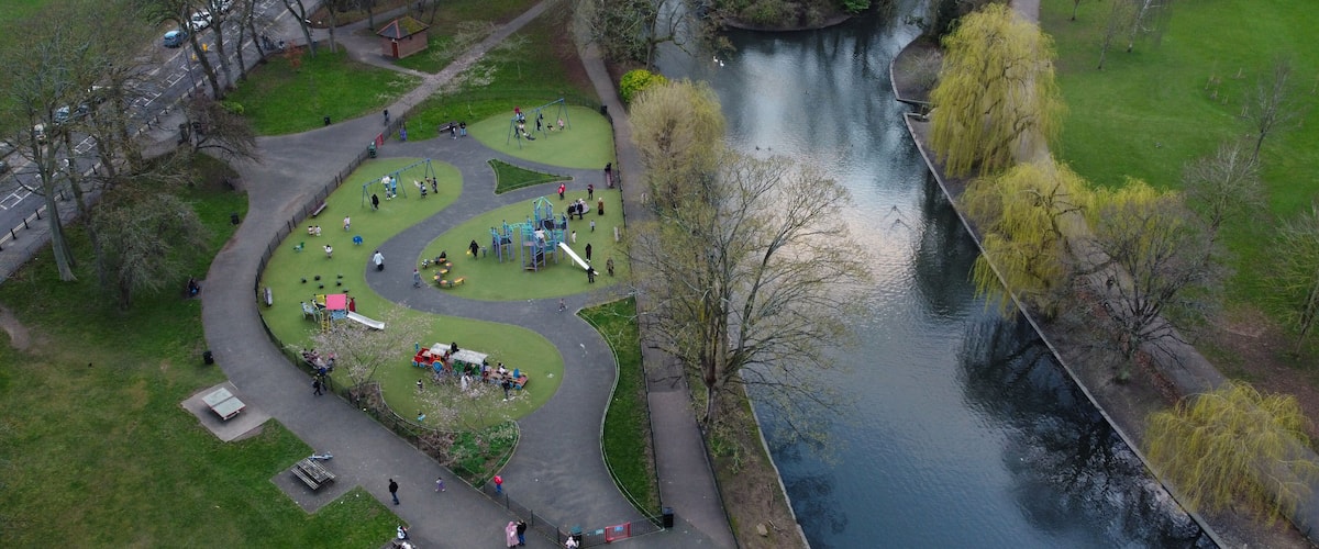An aerial view of a park with a winding path, playgrounds, a river, and a bridge.