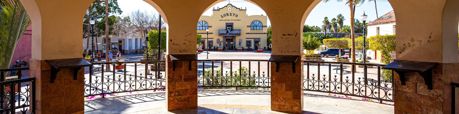 Una plaza con tres arcos, un edificio con la inscripción 'Loretto', y una calle con coches y palmeras.