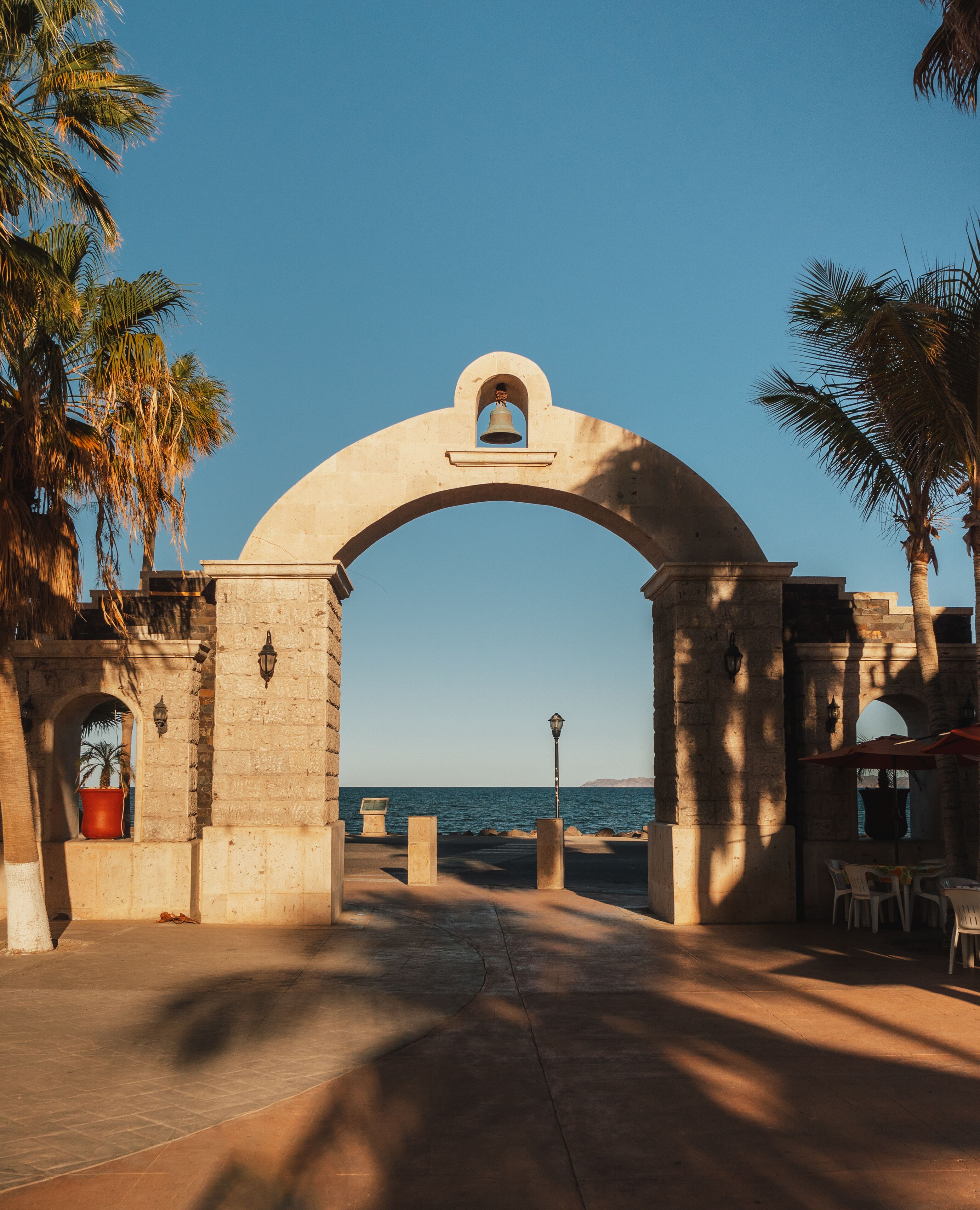 Un arco de piedra con una campana, palmeras y vistas al mar.