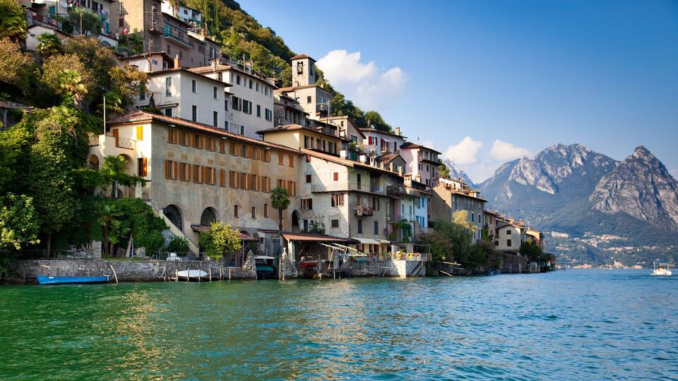 A coastal village with terracotta-roofed buildings, a boat docked on the water, and a mountain in the background.