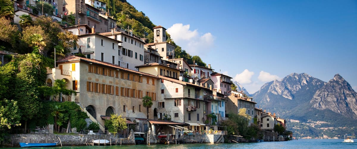A coastal village with terracotta-roofed buildings, a boat docked on the water, and a mountain in the background.