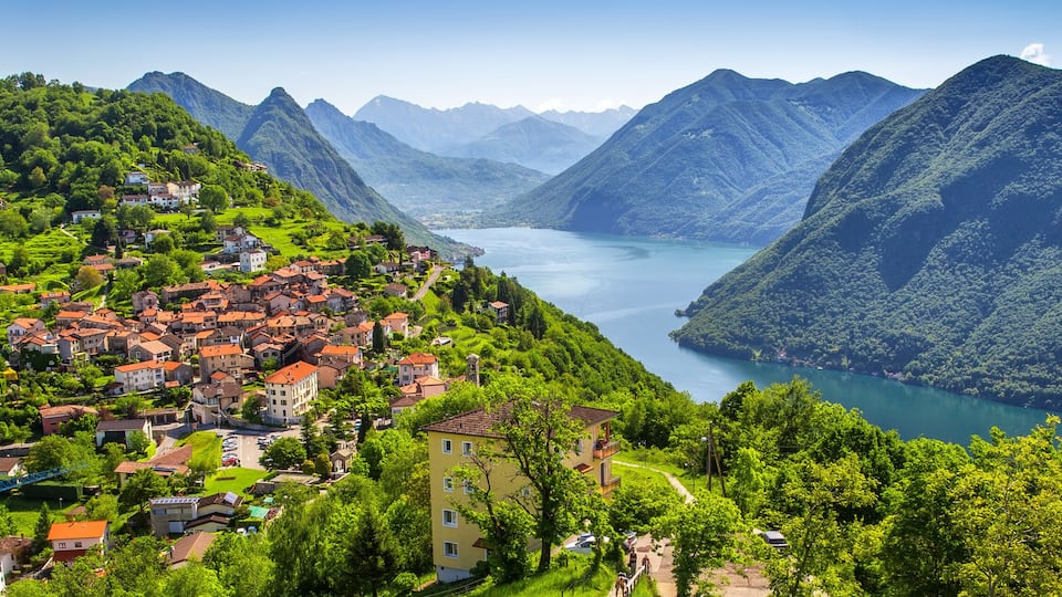 A mountainous village with a lake surrounded by lush greenery and red-roofed houses.