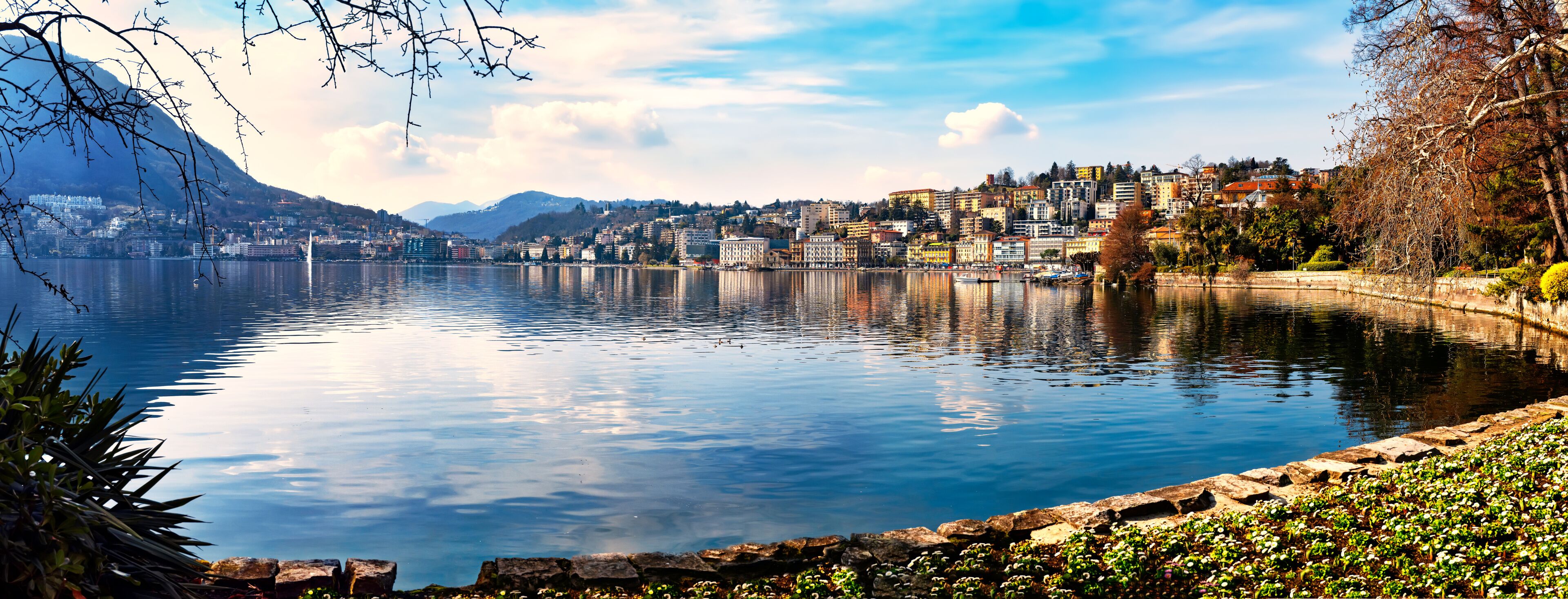 Um lago calmo com um horizonte urbano ao fundo, incluindo montanhas e diversos edifícios.
