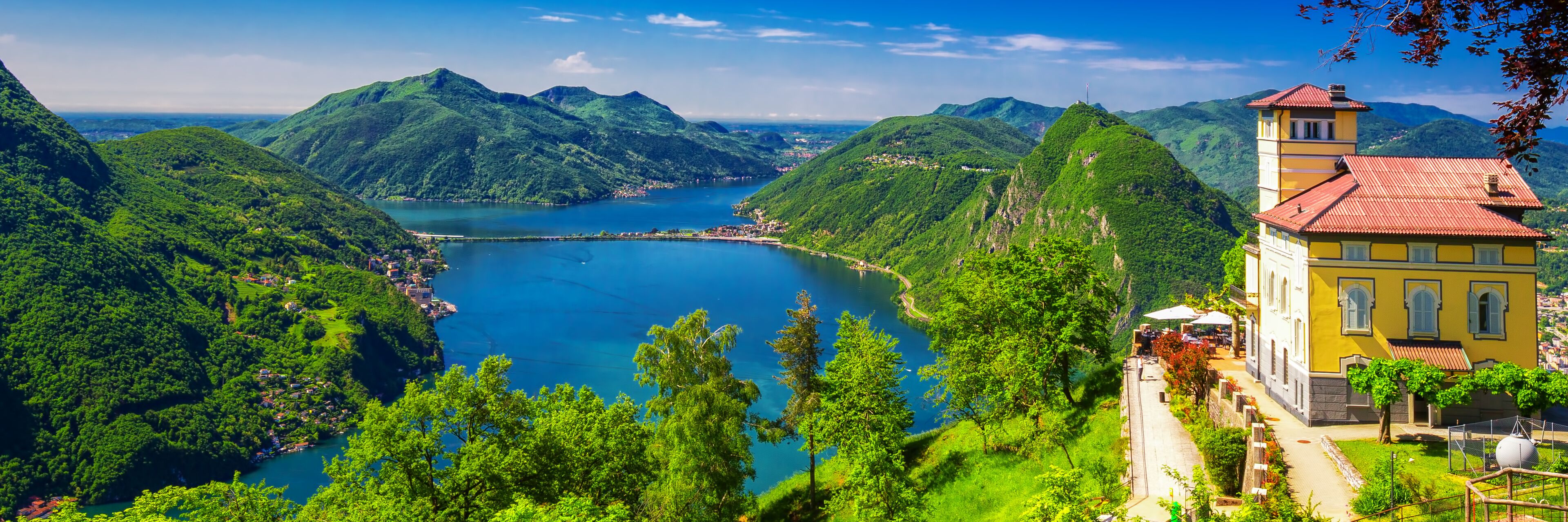 A lake surrounded by lush green hills and a yellow building with a red roof.