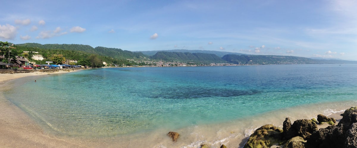 A coastal beach with clear turquoise waters, rocky shoreline, and a town in the background.