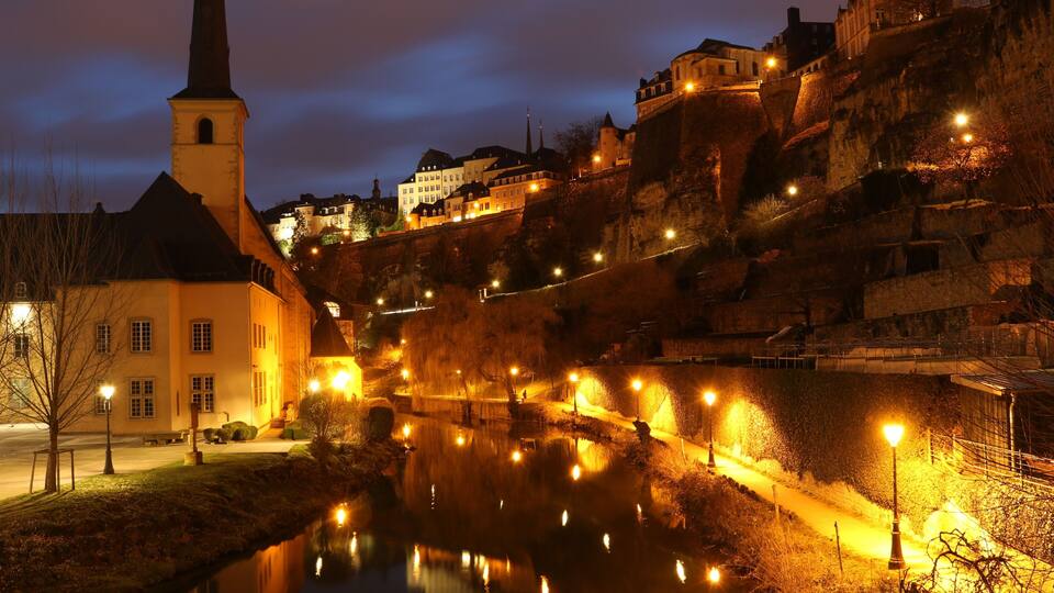 A river reflecting lights, a church with a tall steeple, and buildings on a hillside.