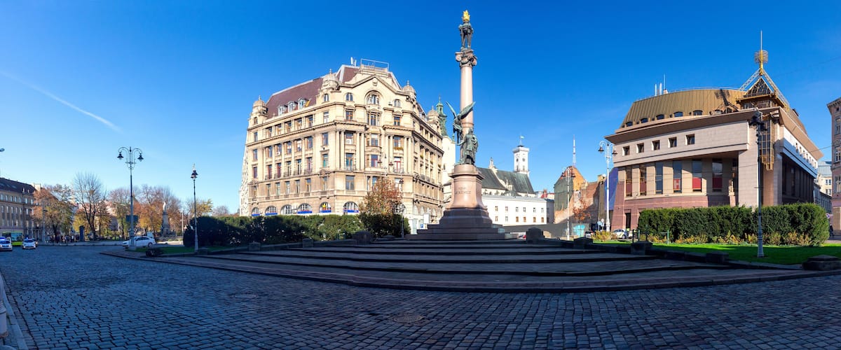 A cobblestone square with a monument, historic buildings, and a clear blue sky.