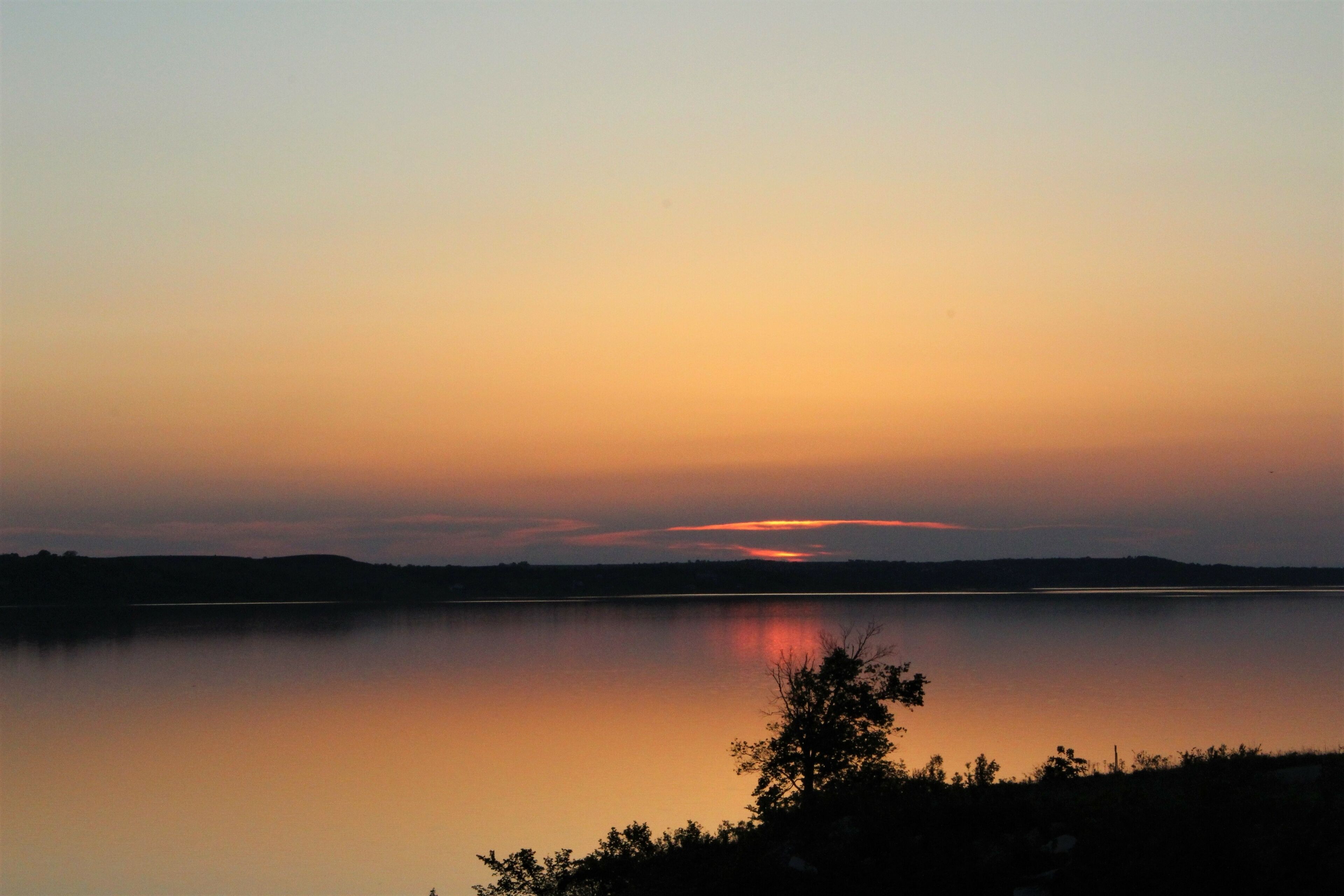 A sunset over a calm lake with silhouettes of trees and distant hills.
