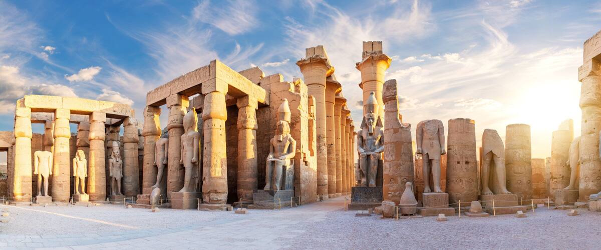 Gamle egyptiske ruiner med høje stensøjler og statuer under en klar himmel.