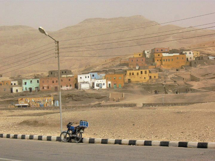Un paisaje rural con un camino de tierra, una motocicleta y edificios coloridos al fondo.
