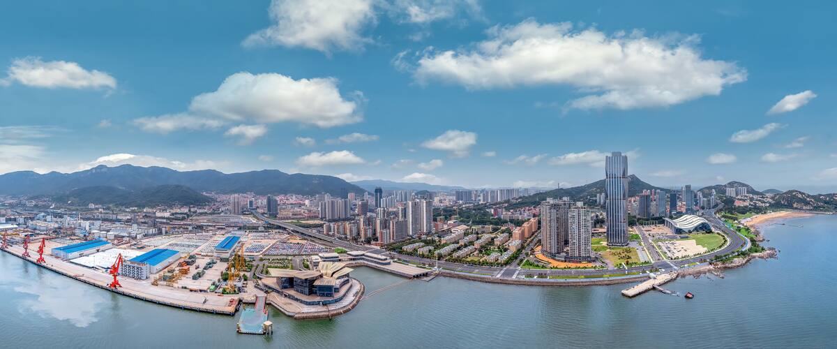 A coastal cityscape with a port, high-rise buildings, and mountains in the background.