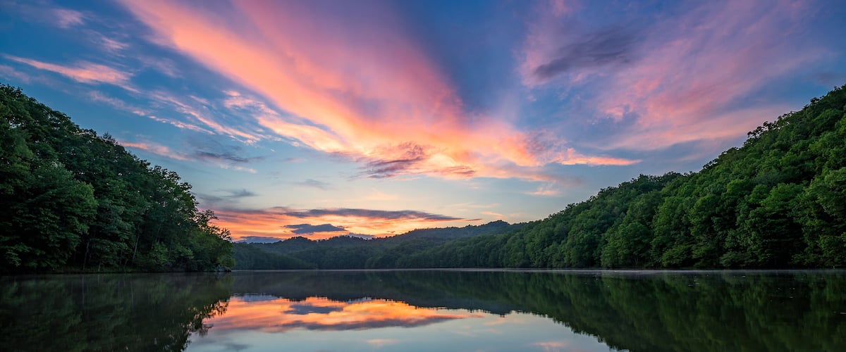 A calm lake with a reflection of the sky, surrounded by dense forest.