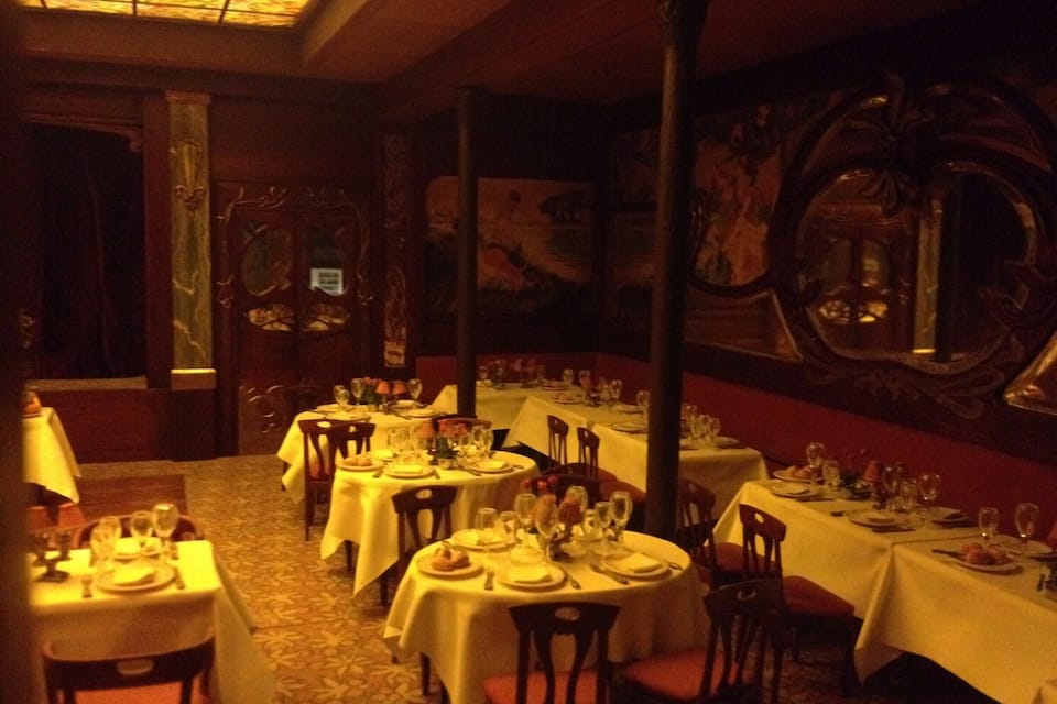A dimly lit restaurant with round tables set for dining, featuring ornate wall decorations and a patterned carpet.