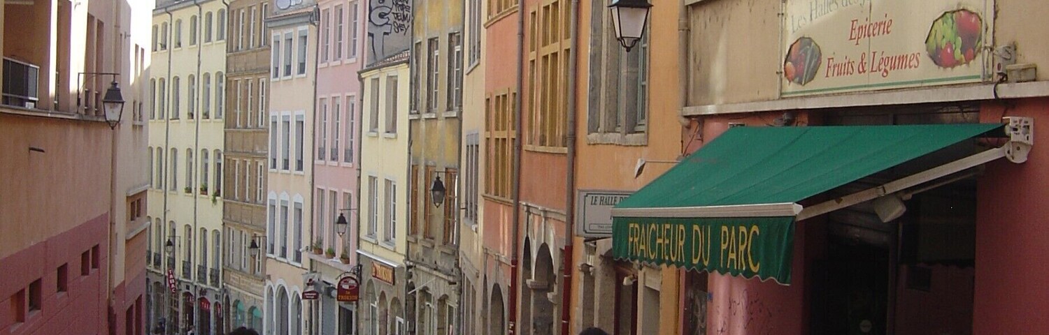 Rua de paralelepípedos flanqueada por edifícios coloridos, um mercado de frutas e legumes, e pedestres a passear.