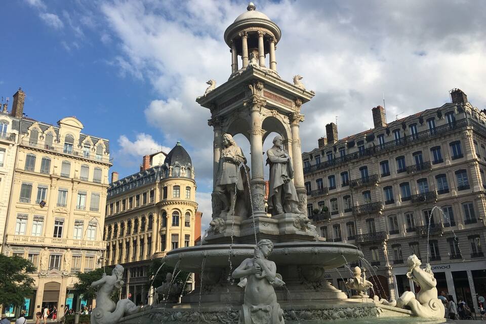 A classical fountain with statues and a central column, surrounded by historic buildings.