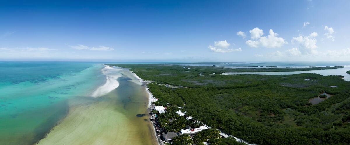 A coastal landscape with a sandy beach, clear turquoise waters, and dense green vegetation.