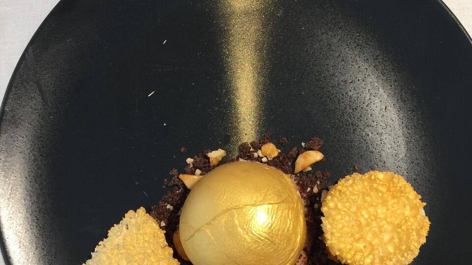 A dessert on a black plate with a golden sphere in the center and four golden meringue pieces around it.