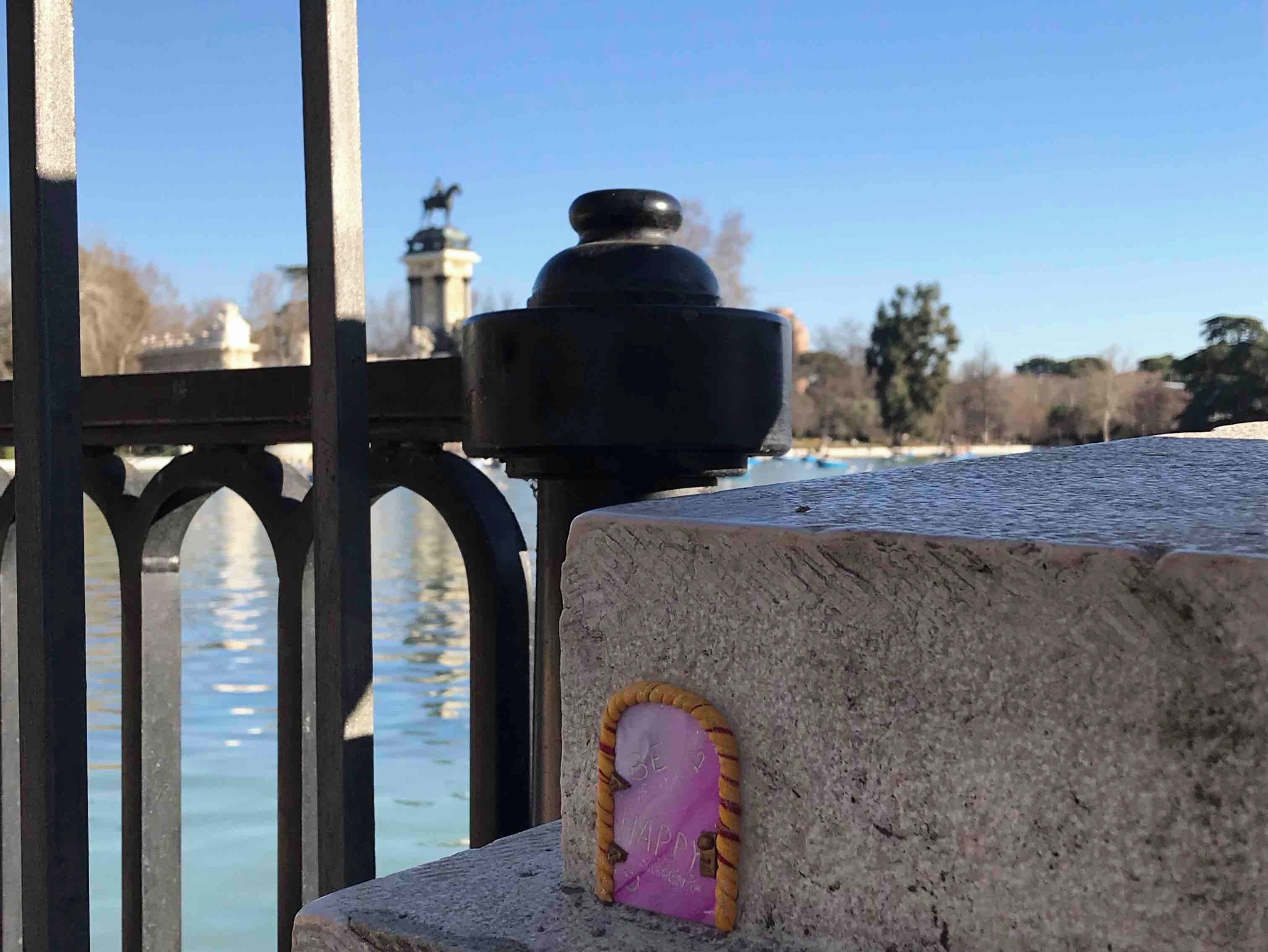 Una estructura de piedra con un detalle decorativo rosa y amarillo, un objeto negro encima y una vista de un río y una estatua distante.