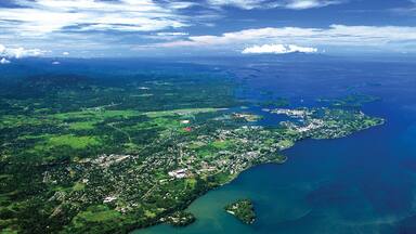An aerial view of a coastal region with dense greenery, a large body of water, and scattered buildings.