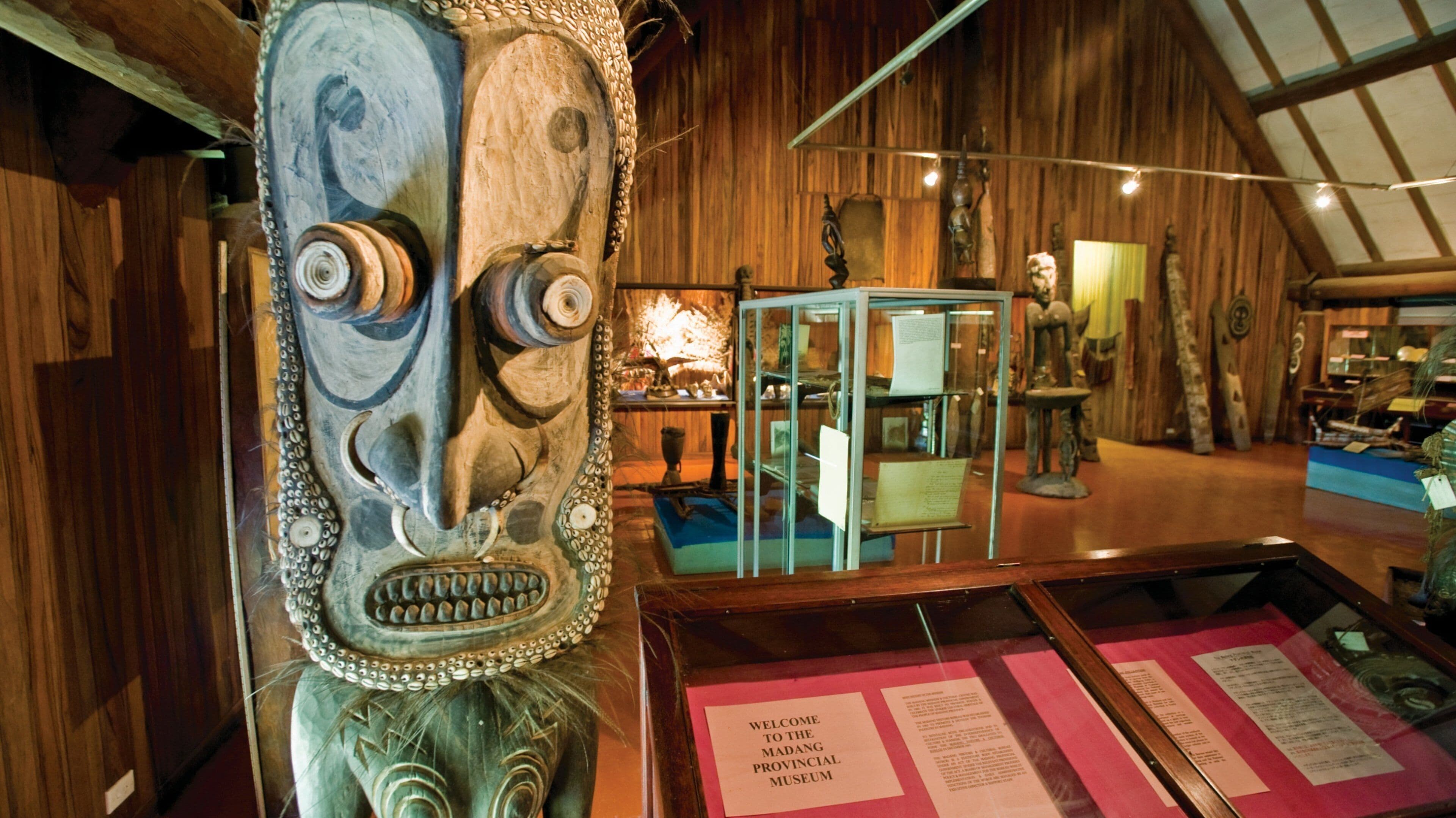 Interior de museo con paredes de madera, una vitrina de cristal y una máscara de madera tallada de gran tamaño.