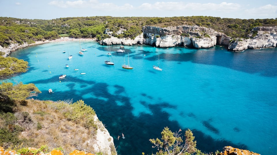 A coastal bay with clear turquoise waters, rocky cliffs, and several boats anchored near the shore.