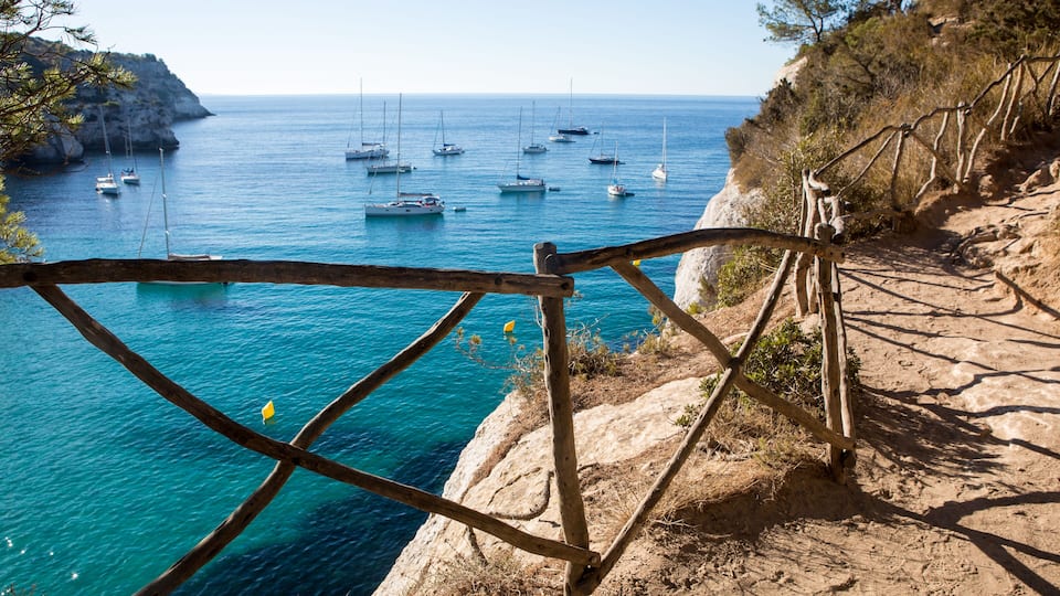 A coastal path with a wooden railing overlooking a turquoise sea with sailboats.