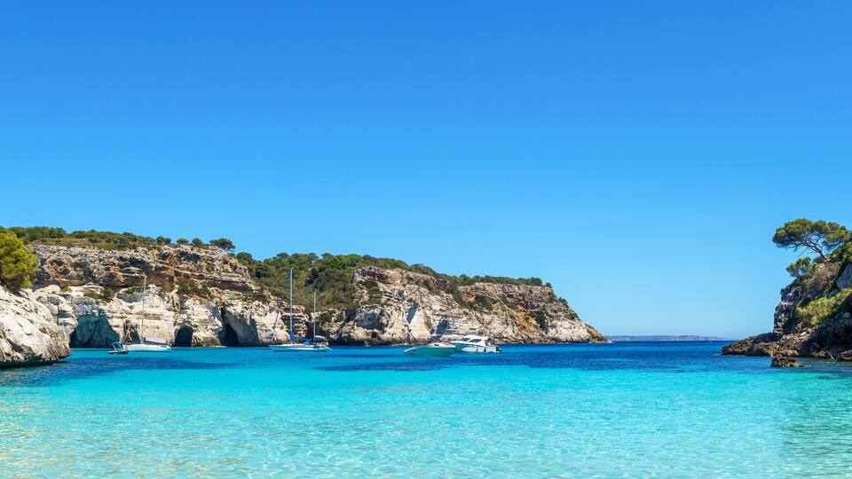 A coastal bay with clear turquoise waters, rocky cliffs, and a sailboat anchored near the shore.