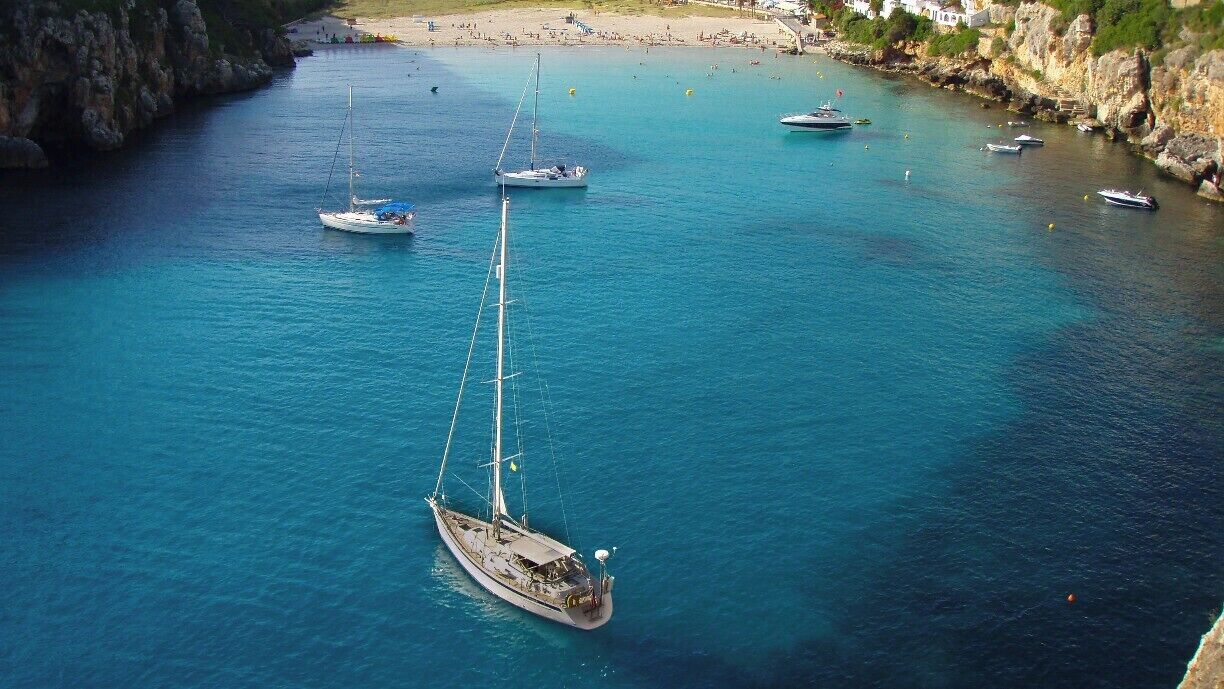 A coastal area with clear turquoise waters, several sailboats, and a rocky shoreline with buildings in the background.