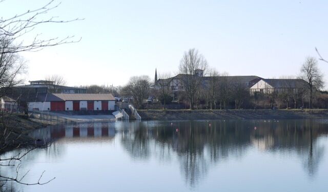 Un lago tranquilo con un edificio rojo y blanco a la izquierda y otro edificio más grande al otro lado del agua.