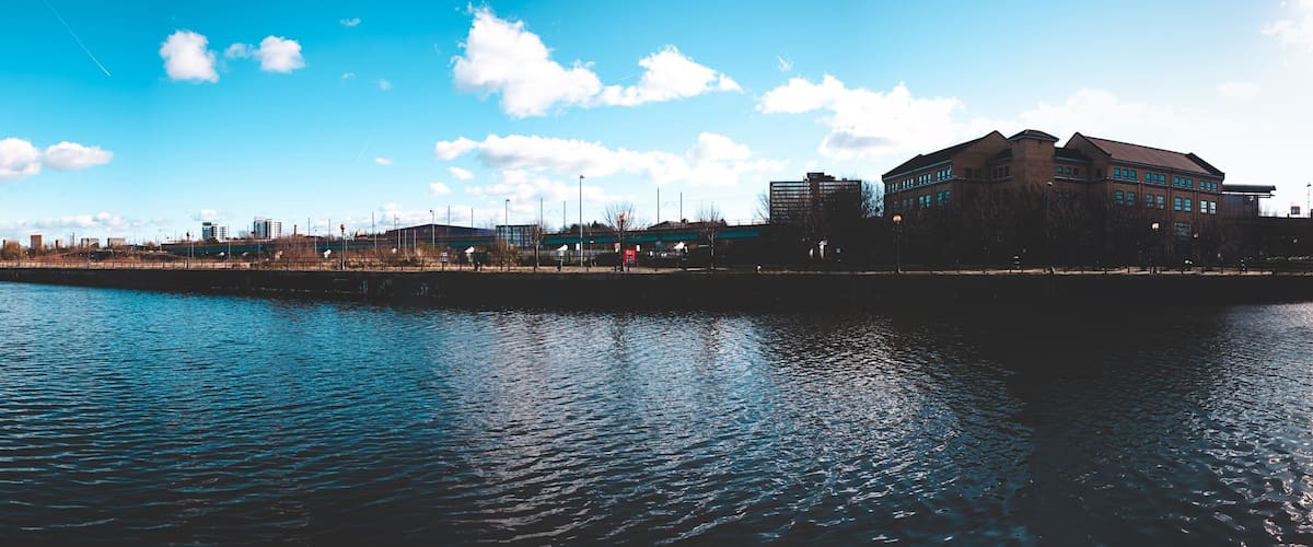 A waterfront cityscape with a prominent building on the right, a calm river, and a clear blue sky.