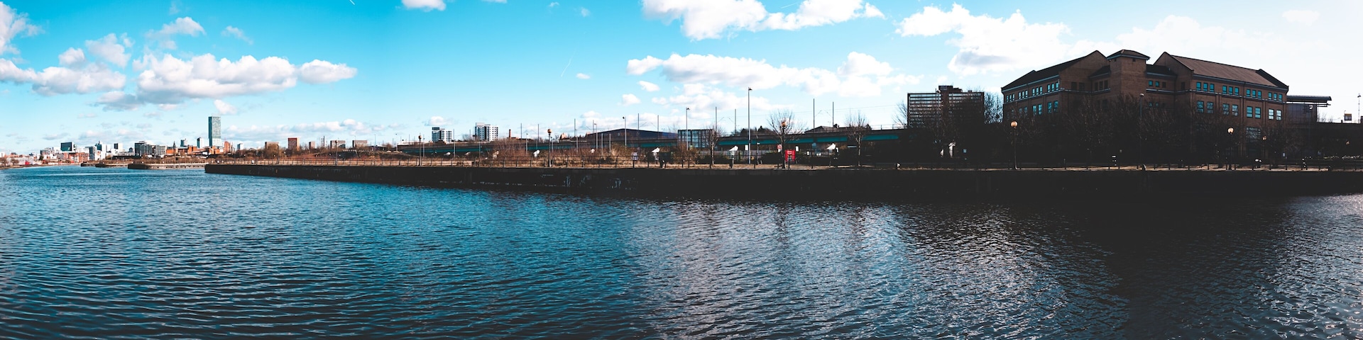 A waterfront cityscape with a prominent building on the right, a calm river, and a clear blue sky.
