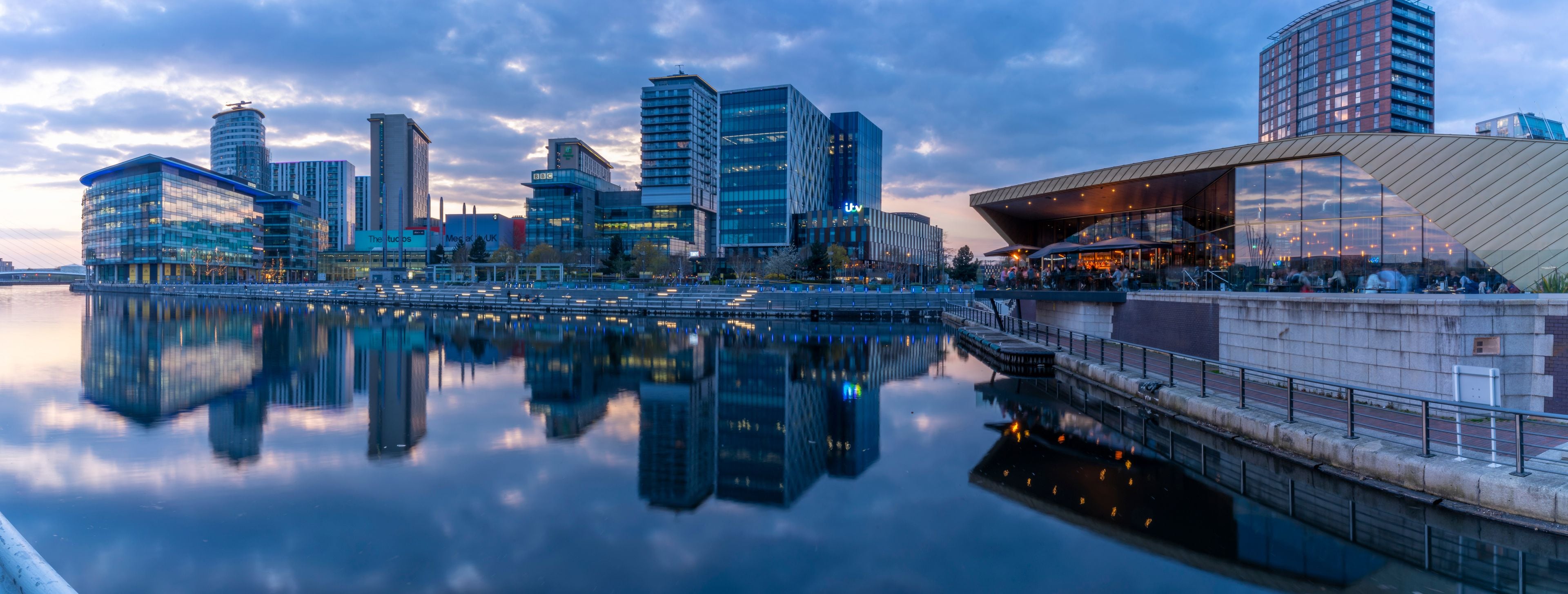 A cityscape with modern buildings, a river reflecting the structures, and a bridge connecting two parts of the city.