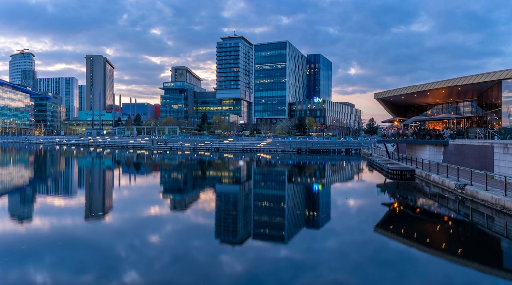 A cityscape with modern buildings, a river reflecting the structures, and a bridge connecting two parts of the city.