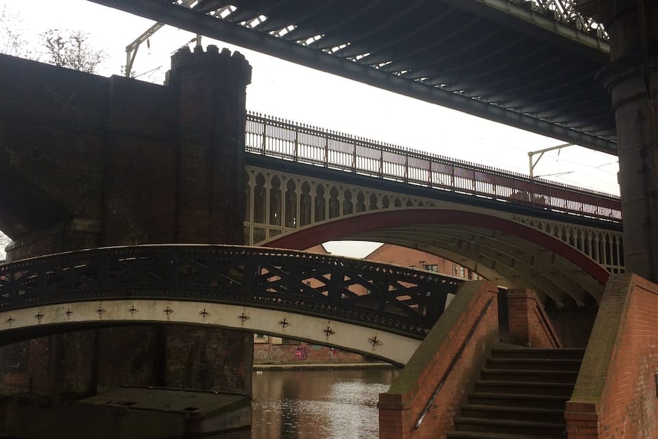 Un puente con diseño de arco gótico, una escalera que lo conduce y un cuerpo de agua debajo.