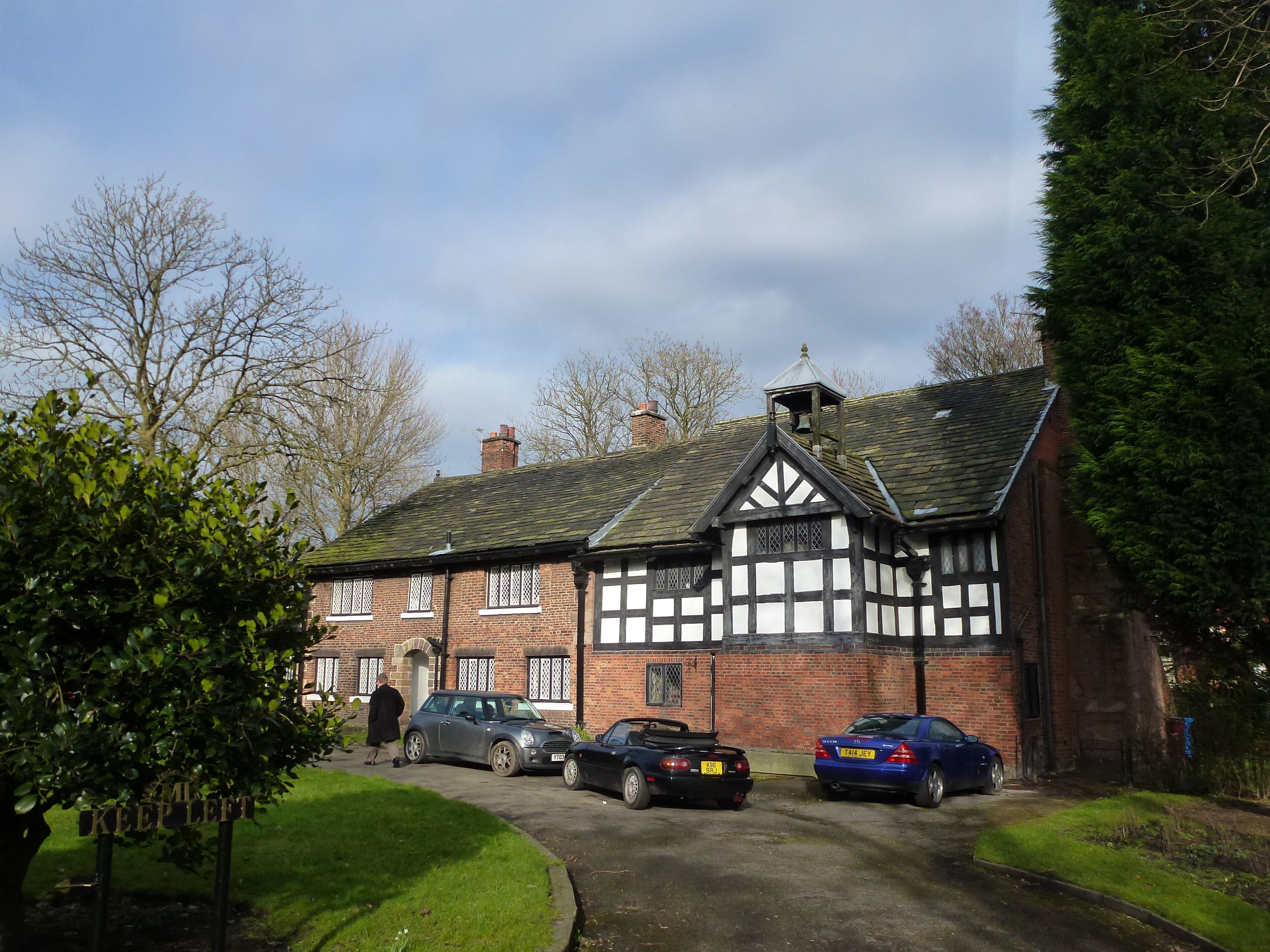 A Tudor-style house with a garden, parked cars, and a 'KEEP LEFT' sign.