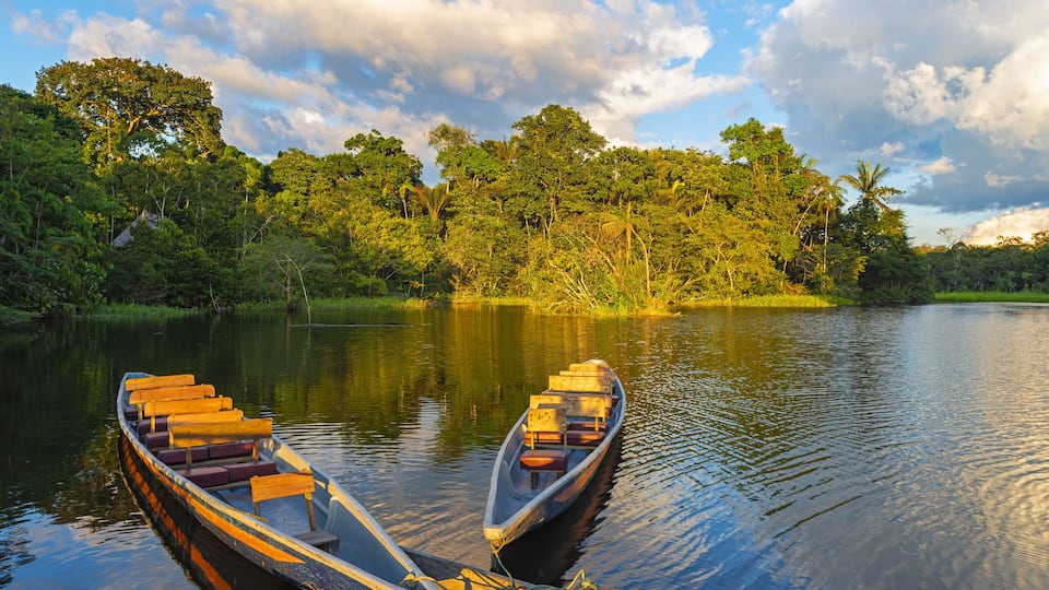 Two wooden boats tied to a dock with a calm river and dense forest in the background.